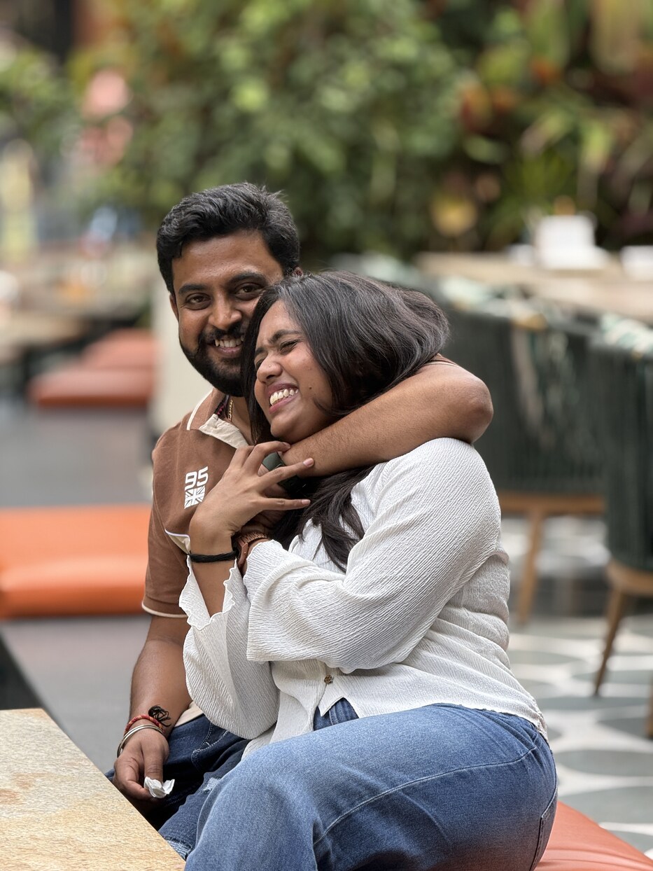 Subramanya hugging Navya from behind at a cafe table, both laughing
