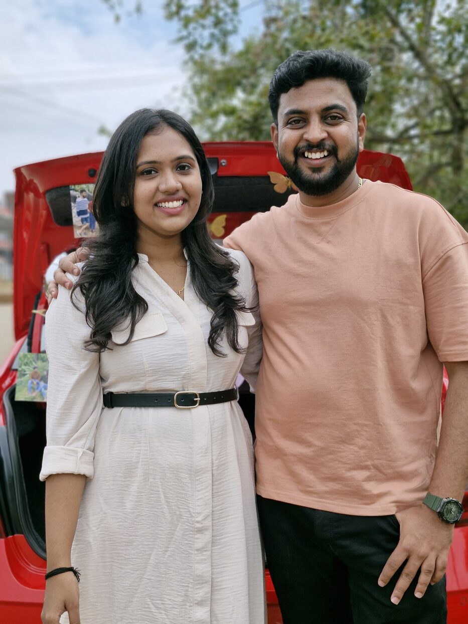 Navya and Subramanya standing by a red car, smiling